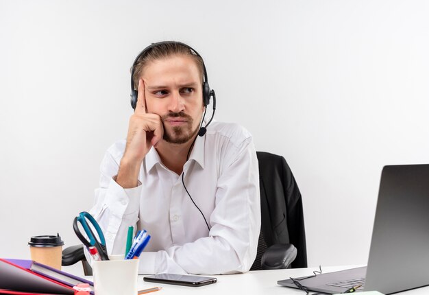 Uomo d'affari bello infastidito in camicia bianca e cuffie con un microfono che ascolta un cliente con la faccia seria che si siede al tavolo in offise sopra priorità bassa bianca