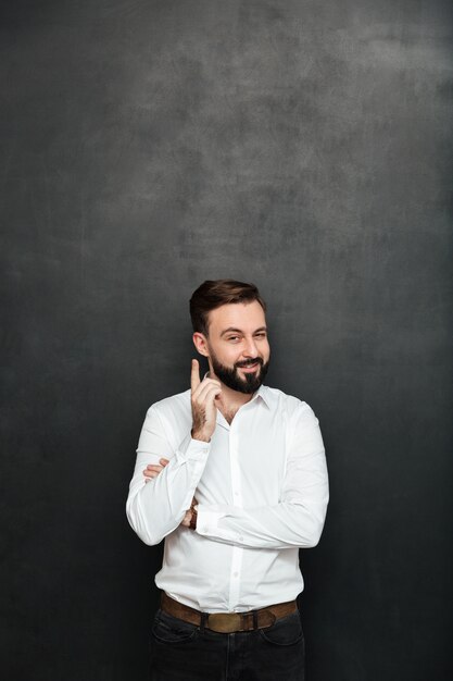 Uomo d'affari castana che posa sulla macchina fotografica con lo sguardo felice ingannevole, indicando il dito indice come se sappia qualcosa sopra grigio scuro