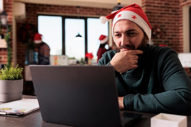 Uomo d'affari che lavora al computer portatile in ufficio decorato con albero di natale e ornamenti festivi. Lavoratore maschio che utilizza il pc sul posto di lavoro con decorazioni natalizie durante la stagione delle vacanze invernali.