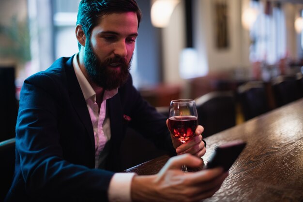 Uomo d'affari che per mezzo del telefono cellulare con bicchiere di vino rosso a disposizione