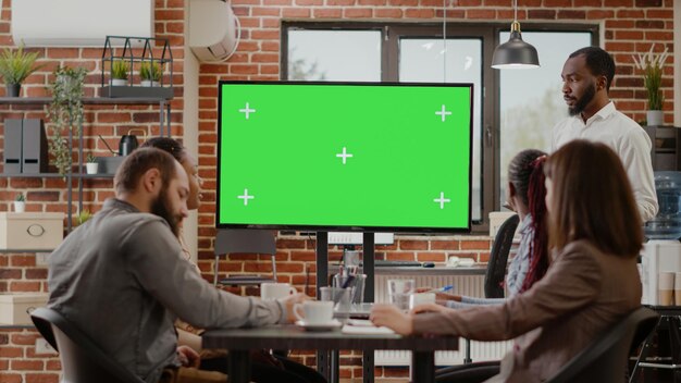 Uomo d'affari che presenta monitor con schermo verde all'incontro con i colleghi, guardando finto sfondo con modello di spazio copia isolato. Persone che lavorano con display chroma key vuoto.