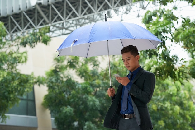 Uomo d'affari cinese che sta con l'ombrello in pioggia e che per mezzo dello smartphone