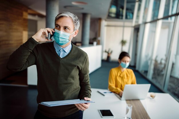 Uomo d'affari con maschera facciale che effettua una telefonata durante l'analisi dei rapporti in ufficio