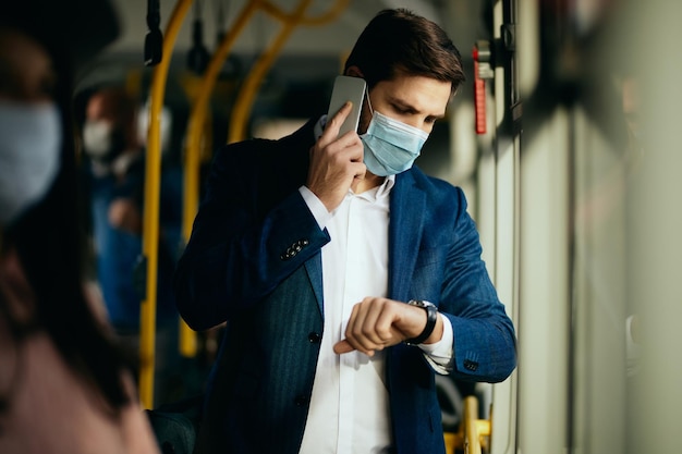 Uomo d'affari con maschera facciale che parla al telefono mentre controlla il tempo sull'orologio da polso in un autobus