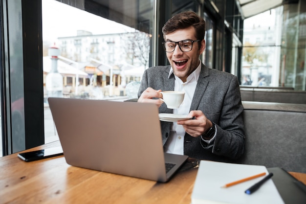 Uomo d'affari di risata in occhiali che si siedono dalla tavola in caffè con la tazza di caffè mentre esaminando computer portatile