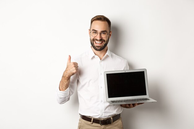 Uomo d'affari di successo che mostra lo schermo del laptop, compone il pollice in segno di approvazione, elogia qualcosa di buono, in piedi