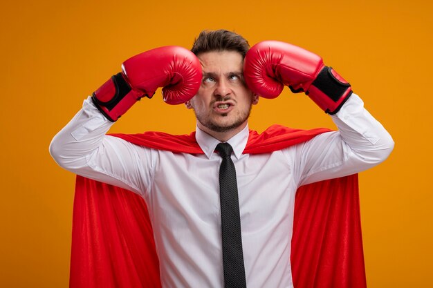 Uomo d'affari dispiaciuto del supereroe in mantello rosso e guantoni da boxe che si prende a pugni alla testa facendo una smorfia in piedi sopra il muro arancione