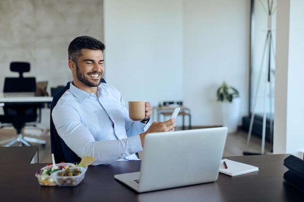 Uomo d'affari felice che manda un sms sullo smartphone mentre beve caffè in ufficio