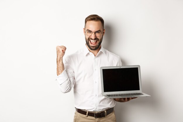 Uomo d'affari felice che mostra lo schermo del laptop, fa la pompa del pugno e si rallegra per i risultati online, in piedi