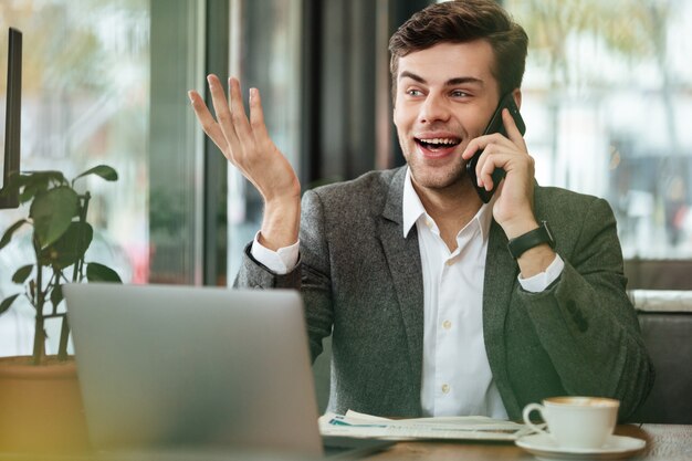 Uomo d'affari felice che si siede dalla tavola in caffè con il computer portatile e che parla dallo smartphone mentre distogliendo lo sguardo