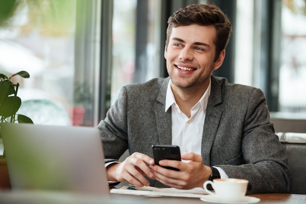 Uomo d'affari felice che si siede dalla tavola in caffè con il computer portatile e lo smartphone mentre distogliendo lo sguardo