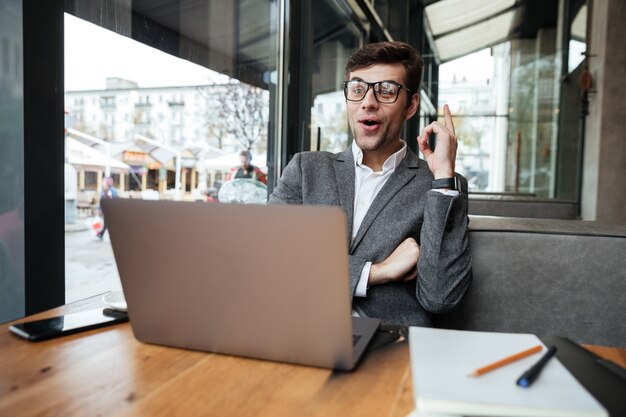 Uomo d'affari felice in occhiali che si siedono dalla tavola in caffè con il computer portatile e che hanno idea