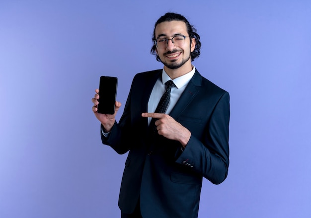Uomo d'affari in abito nero e occhiali che mostra lo smartphone che punta con il dito ad esso sorridente e ammiccante in piedi sopra la parete blu