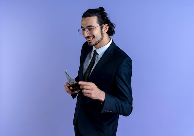 Uomo d'affari in abito nero e occhiali che mostrano contanti e carta di credito sorridendo allegramente in piedi sopra la parete blu