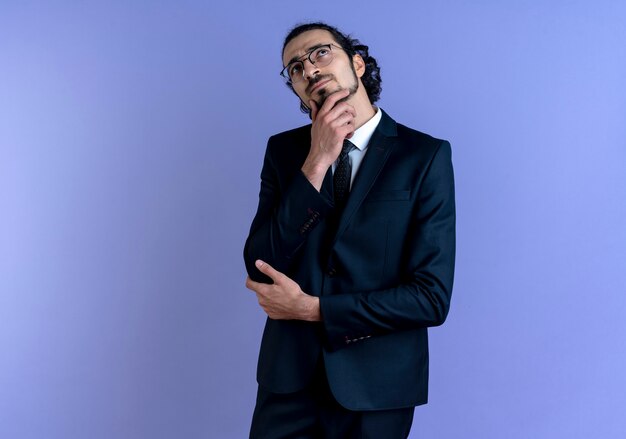 Uomo d'affari in abito nero e occhiali che osserva in su con la mano sul mento perplesso in piedi sopra la parete blu