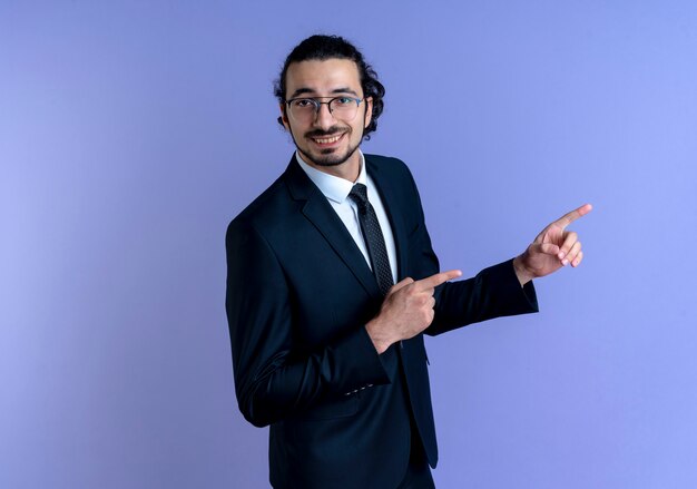 Uomo d'affari in abito nero e occhiali che punta con il dito indice a lato sorridendo allegramente in piedi sopra la parete blu