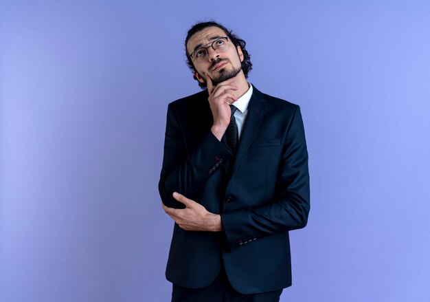 Uomo d'affari in abito nero e occhiali guardando con espressione pensierosa sul viso in piedi sopra la parete blu
