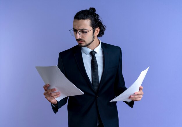 Uomo d'affari in abito nero e occhiali guardando i documenti con la faccia seria in piedi sopra la parete blu