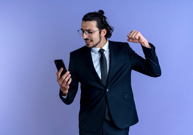 Uomo d'affari in abito nero e occhiali guardando il suo smartphone alzando il pugno in piedi eccitato e arrabbiato sopra la parete blu
