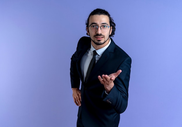 Uomo d'affari in abito nero e occhiali guardando in avanti con il braccio fuori come fare una domanda in piedi sopra la parete blu