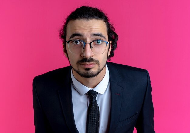 Uomo d'affari in abito nero e occhiali guardando in avanti sorpreso con gli occhi spalancati in piedi sopra il muro rosa