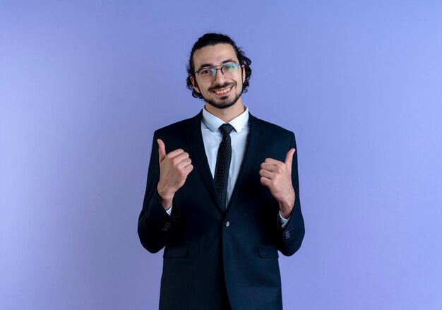 Uomo d'affari in abito nero e occhiali guardando in avanti sorridendo allegramente mostrando i pollici in su in piedi sopra la parete blu