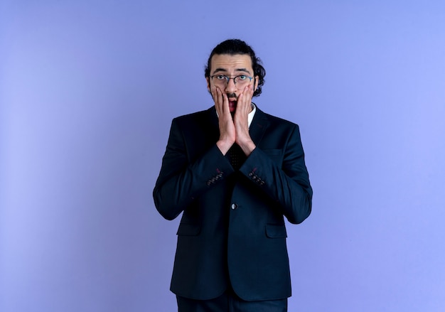 Uomo d'affari in abito nero e occhiali guardando la bocca di copertura scioccata anteriore con la mano in piedi sopra la parete blu