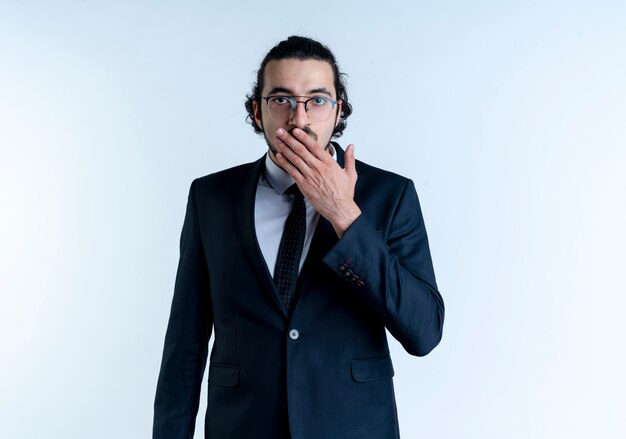Uomo d'affari in abito nero e occhiali guardando la bocca di copertura scioccata anteriore con la mano in piedi sul muro bianco