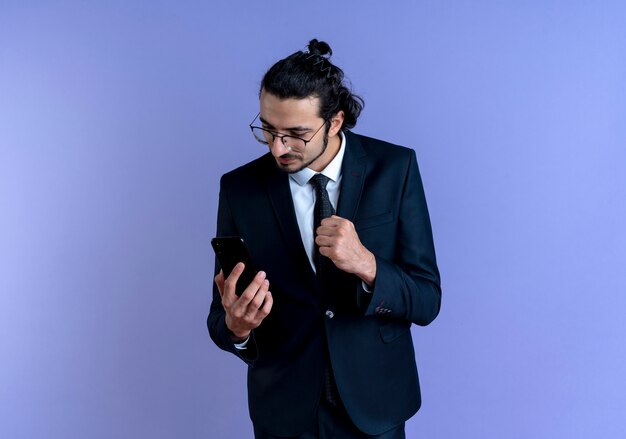 Uomo d'affari in abito nero e occhiali guardando lo schermo del suo smartphone stringendo il pugno in piedi felice e positivo sopra la parete blu