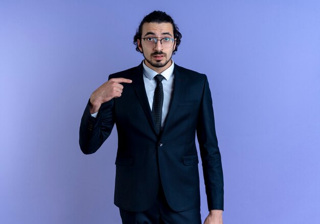 Uomo d'affari in abito nero e occhiali puntati con il dito a se stesso cercando confuso in piedi sopra la parete blu