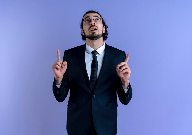Uomo d'affari in abito nero e occhiali rivolti con il dito indice in alto guardando fiducioso in piedi oltre la parete blu