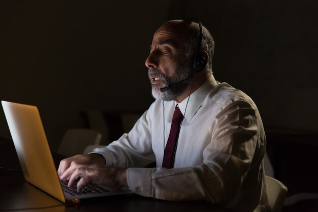 Uomo d'affari in cuffia avricolare che parla e che utilizza computer portatile