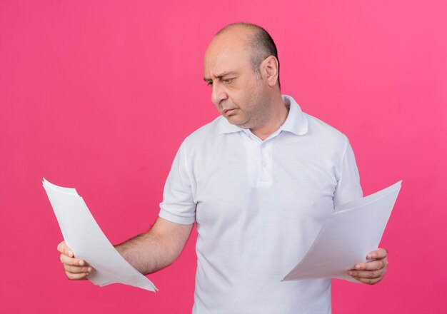 Uomo d'affari maturo casuale concentrato che tiene e che esamina i documenti isolati su fondo rosa