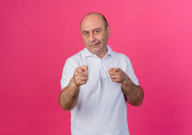 Uomo d'affari maturo casuale sicuro che ti fa gesto alla macchina fotografica isolata su fondo rosa con lo spazio della copia