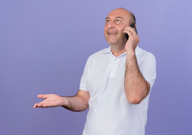 Uomo d'affari maturo casuale sorridente che osserva in su che parla sul telefono e che mostra la mano vuota isolata su fondo viola