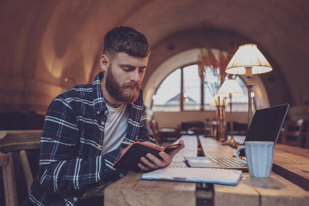 Uomo d'affari occasionale o libero professionista che pianifica il suo lavoro su notebook, lavorando su computer portatile con smartphone, tazza di caffè sul tavolo al bar o ufficio a casa, lavorando dal concetto di caffè