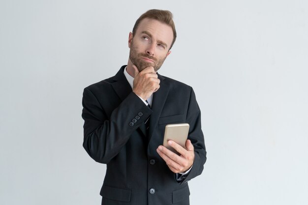 Uomo d&#39;affari pensoso toccando il mento, pensando e tenendo smartphone.