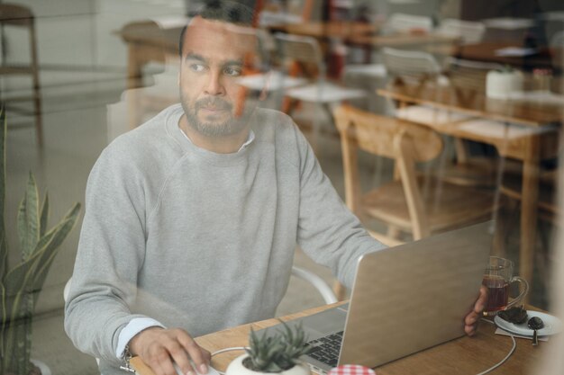 Uomo d'affari premuroso in maglione che guarda intensamente nella finestra durante il lavoro sul laptop nel caffè della città