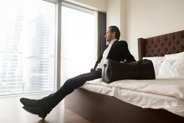 Uomo d&#39;affari rilassato che si siede sul letto oltre alla borsa dei bagagli.
