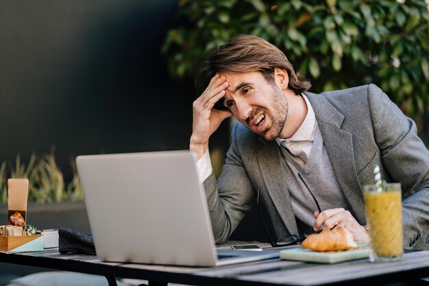 Uomo d'affari scontento che lavora al computer portatile mentre è seduto nella caffetteria all'aperto.