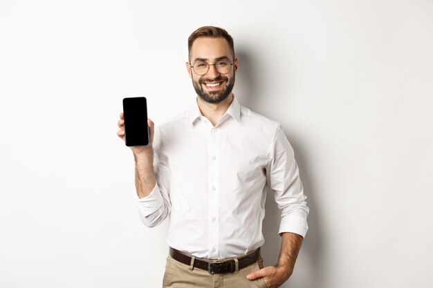 Uomo d'affari soddisfatto che mostra lo schermo mobile, sorridendo con orgoglio, in piedi su sfondo bianco.