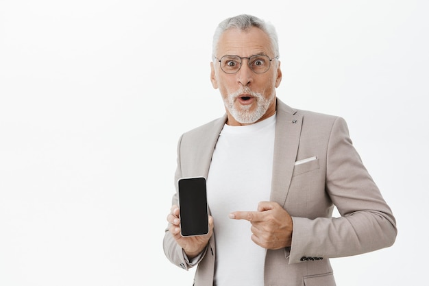 Uomo d'affari sorpreso e stupito in tuta puntare il dito sullo schermo dello smartphone, mostrando l'applicazione