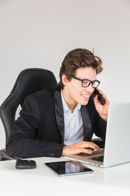 Uomo d&#39;affari sorridente che per mezzo del computer portatile mentre parlando sullo smartphone