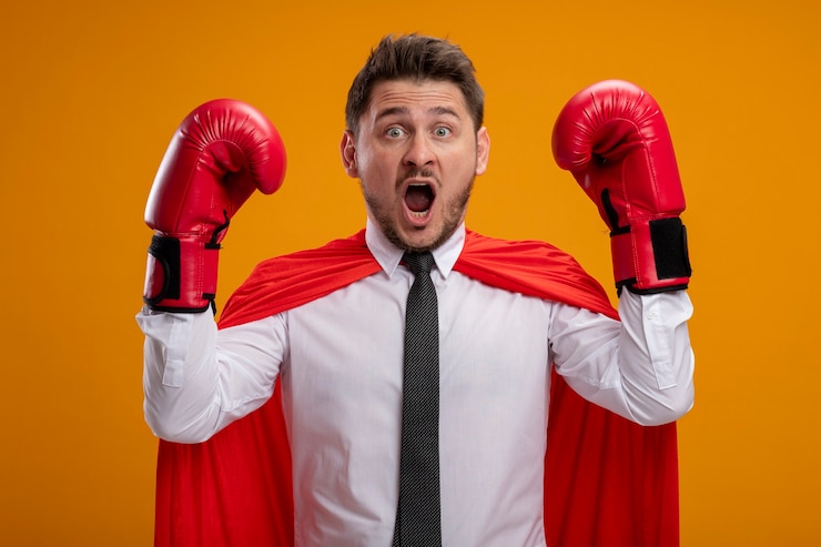 Foto gratuito uomo d'affari super eroe in mantello rosso e guantoni da boxe alzando le mani spaventato gridando in piedi sopra la parete arancione