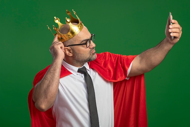 Uomo d'affari super eroe in mantello rosso e occhiali che indossa la corona facendo selfie utilizzando lo smartphone