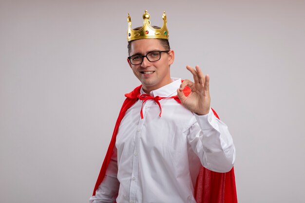 Uomo d'affari super eroe in mantello rosso e occhiali che indossano corona sorridendo allegramente mostrando segno giusto in piedi sopra il muro bianco
