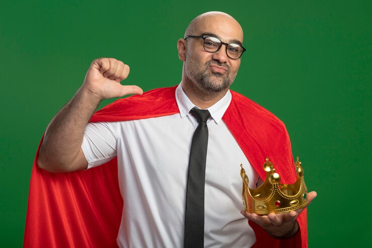 Foto gratuito uomo d'affari super eroe in mantello rosso e occhiali che tengono la corona che punta a se stesso sorridente in piedi sicuro sopra la parete verde
