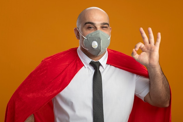 Uomo d'affari super eroe in maschera facciale protettiva e mantello rosso guardando davanti sorridente che mostra segno giusto in piedi sopra la parete arancione