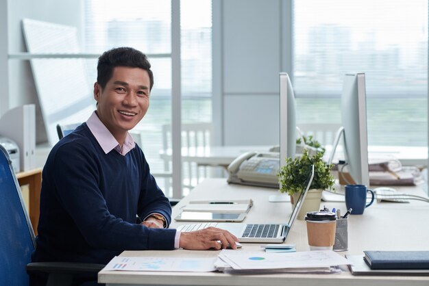 Uomo d'affari vietnamita sorridente che si siede allo scrittorio in ufficio e che esamina macchina fotografica