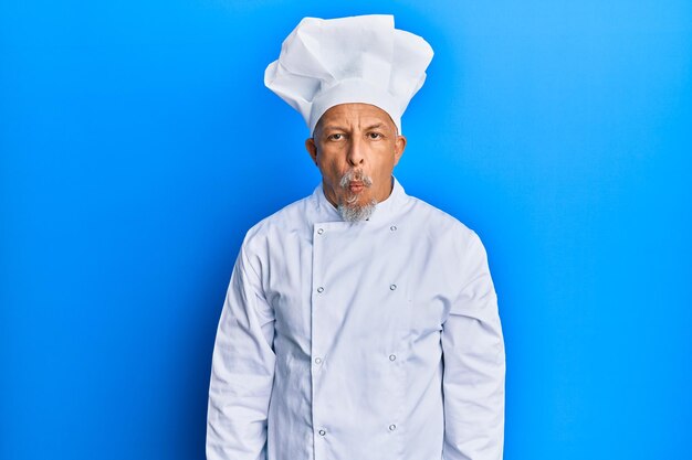 Uomo dai capelli grigi di mezza età che indossa un'uniforme da cuoco professionista e un cappello che fa la faccia da pesce con le labbra, gesto folle e comico. espressione buffa.